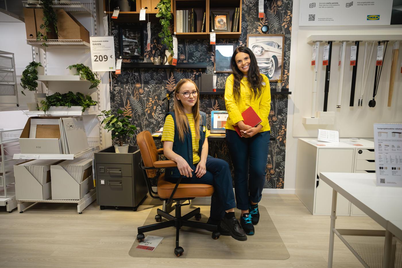 Dvi IKEA ekspertės su geltonos ir mėlynos spalvų uniformomis biuro baldų ekspozicijoje. Viena sėdi biuro kėdėje, kita stovi šalia laikydama aplanką.