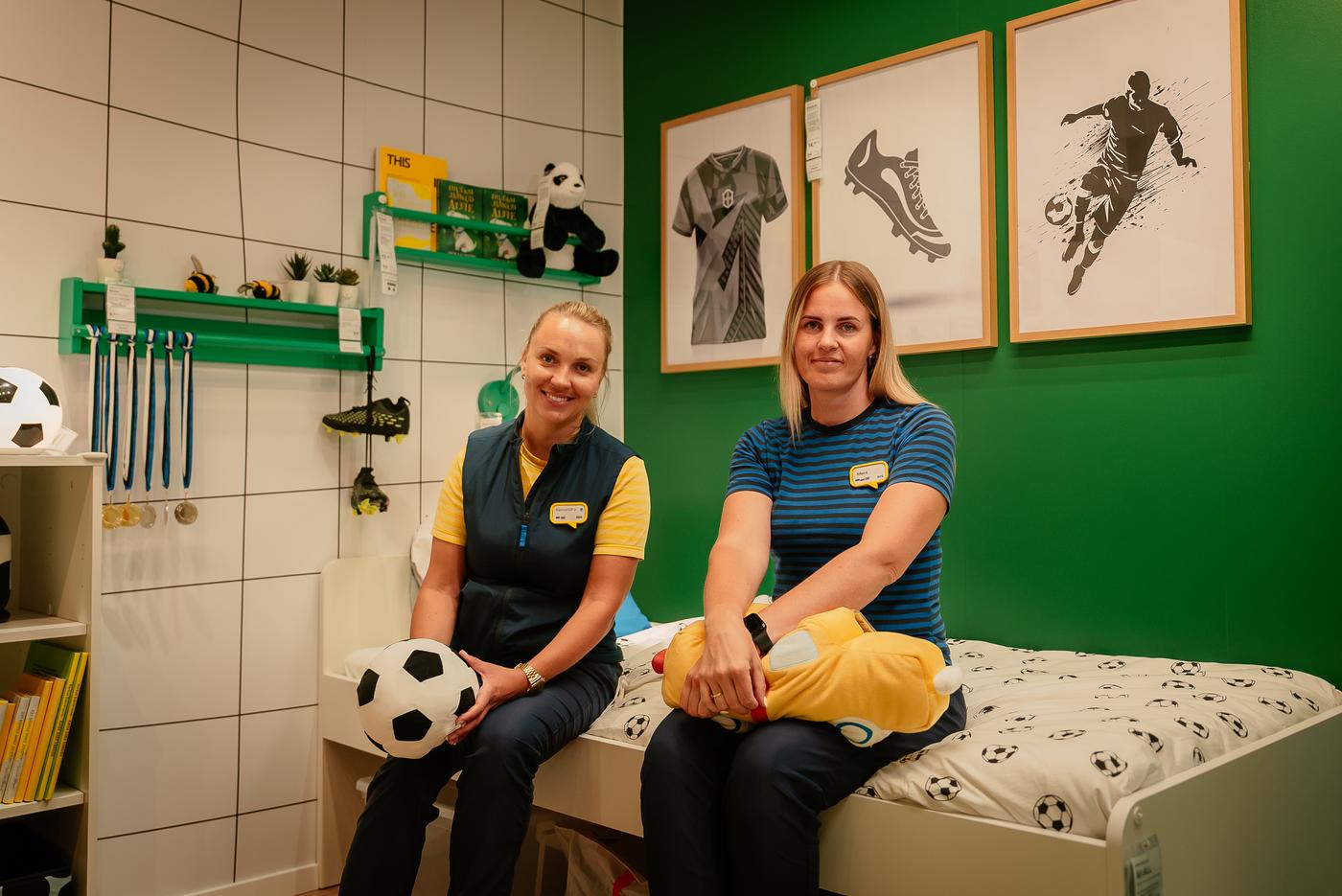 Kaks sinikollases vormis IKEA töötajat istuvad sporditeemaliselt sisustatud lastetoas voodi serval. Ühel neist on käes mängujalgpall ning teisel kollane pehme mänguasi.	