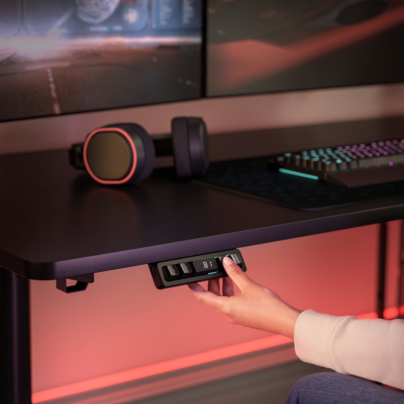 A close-up of a black MÅLOMRÅDE gaming desk. A person’s hand is pressing a button to adjust the height of the desk.