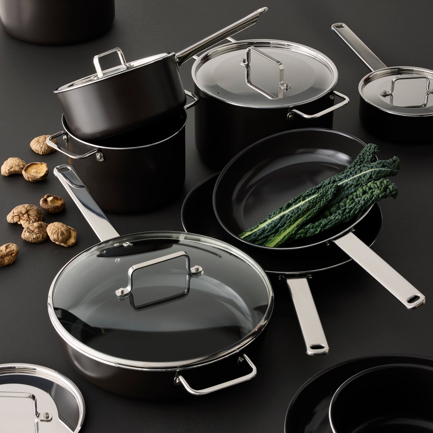 A close-up of a black-grey GULDÖRING sauté pan with lid is placed on a table with greens and nuts nearby.
