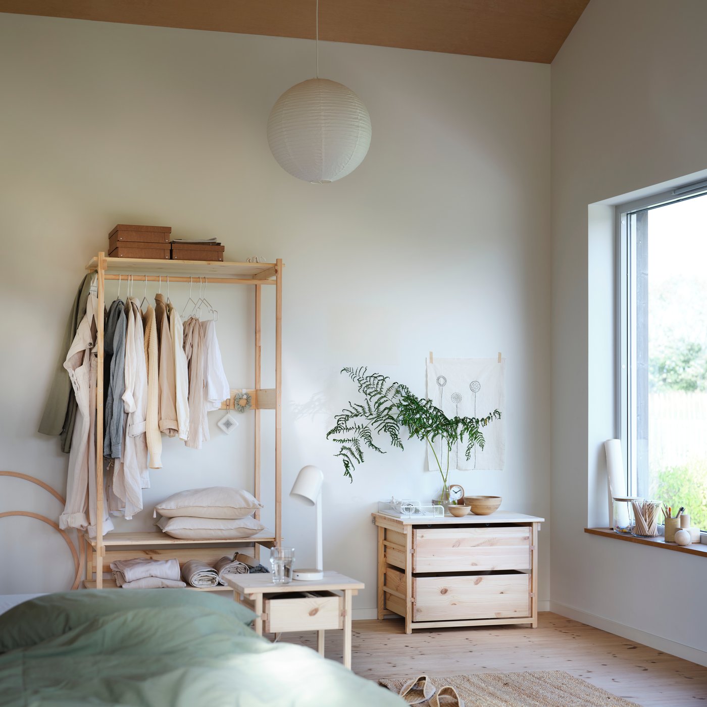A light-filled bedroom with the KRONÖREN storage family. The open wardrobe holds neutral coloured clothing and the chest of drawers holds a plant.