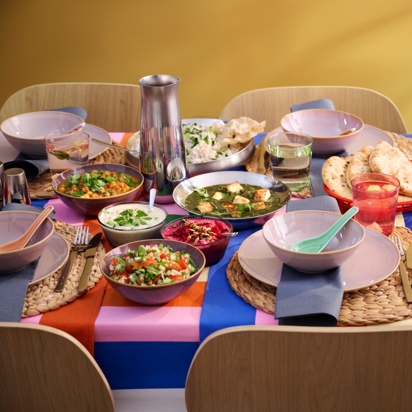 A colourful dining table with bowls of curries, sides, bread, and drinks on a striped tablecloth.